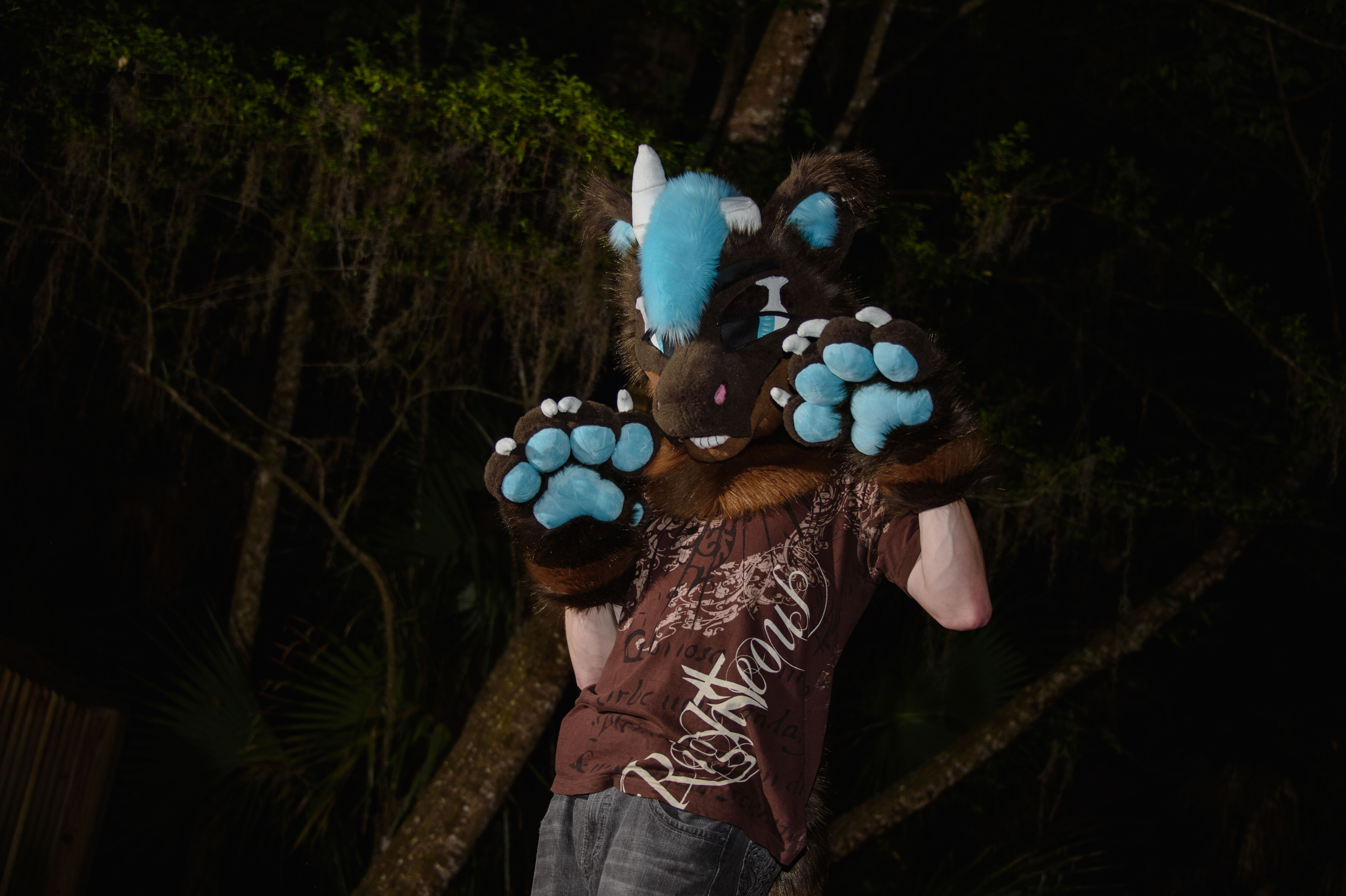 A person in a brown and blue fursuit standing in front of a dark, forested background, with both of their paws in the air