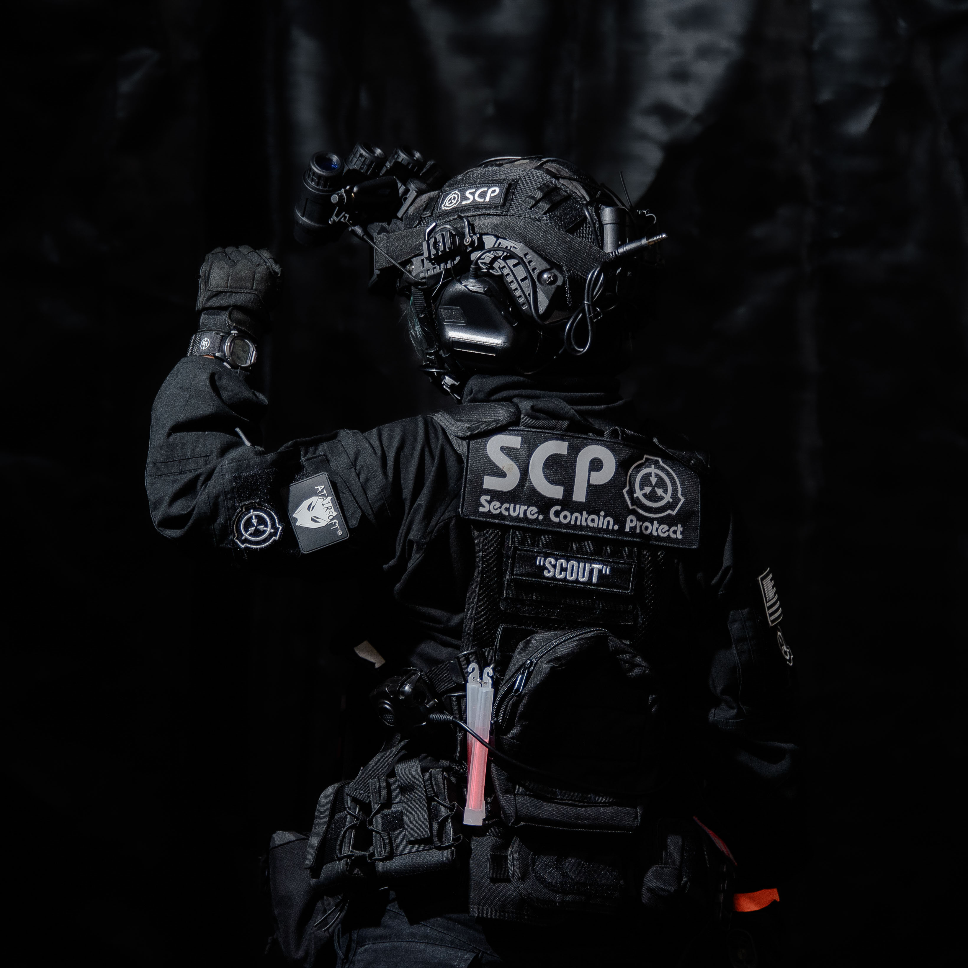 A person in SCP tactical gear, with their back towards the camera, holding their left arm upwards with a fist. They are looking to the left.