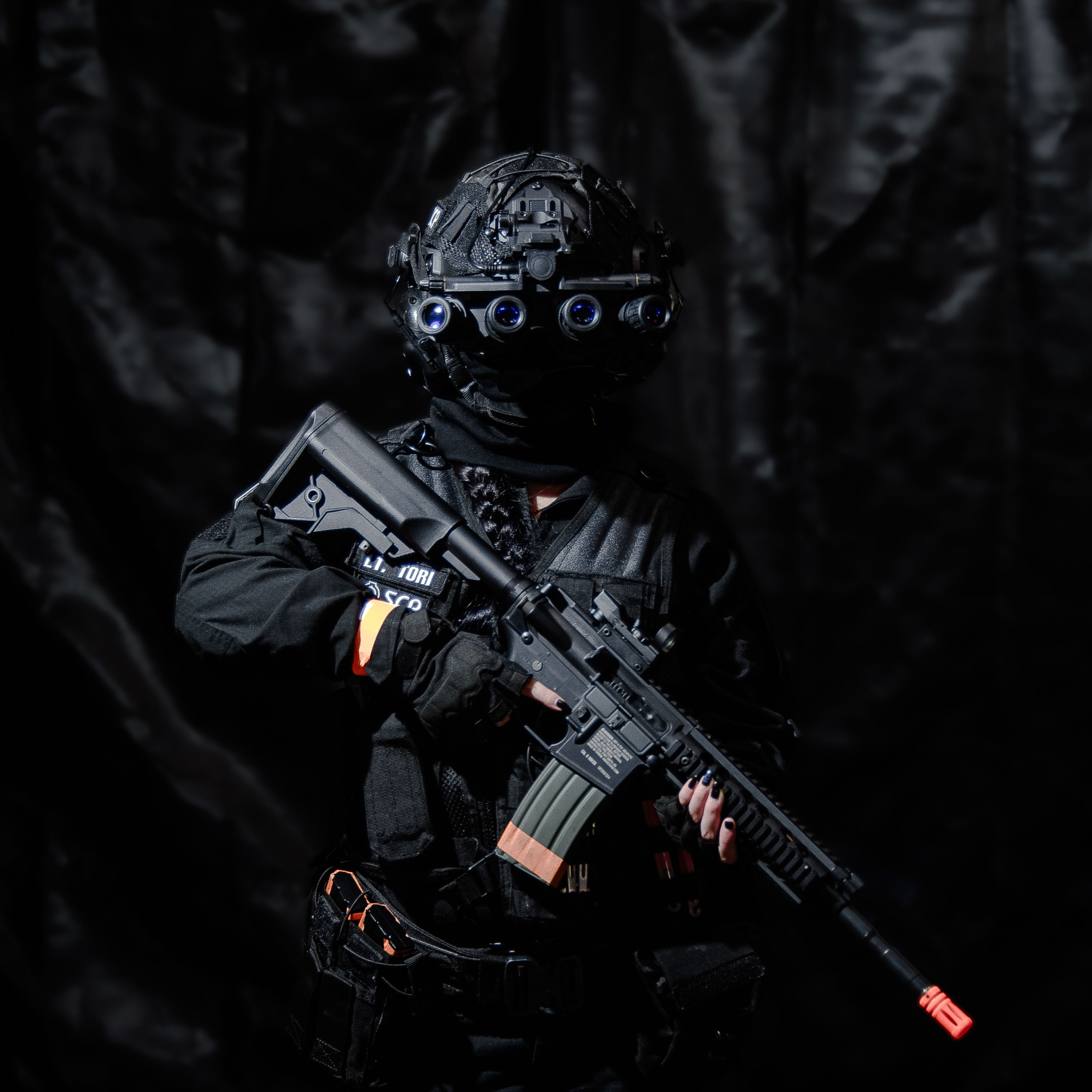 A person in full black tactical gear, in front of a black background, facing towards the camera, holding an assault rifle against their chest.