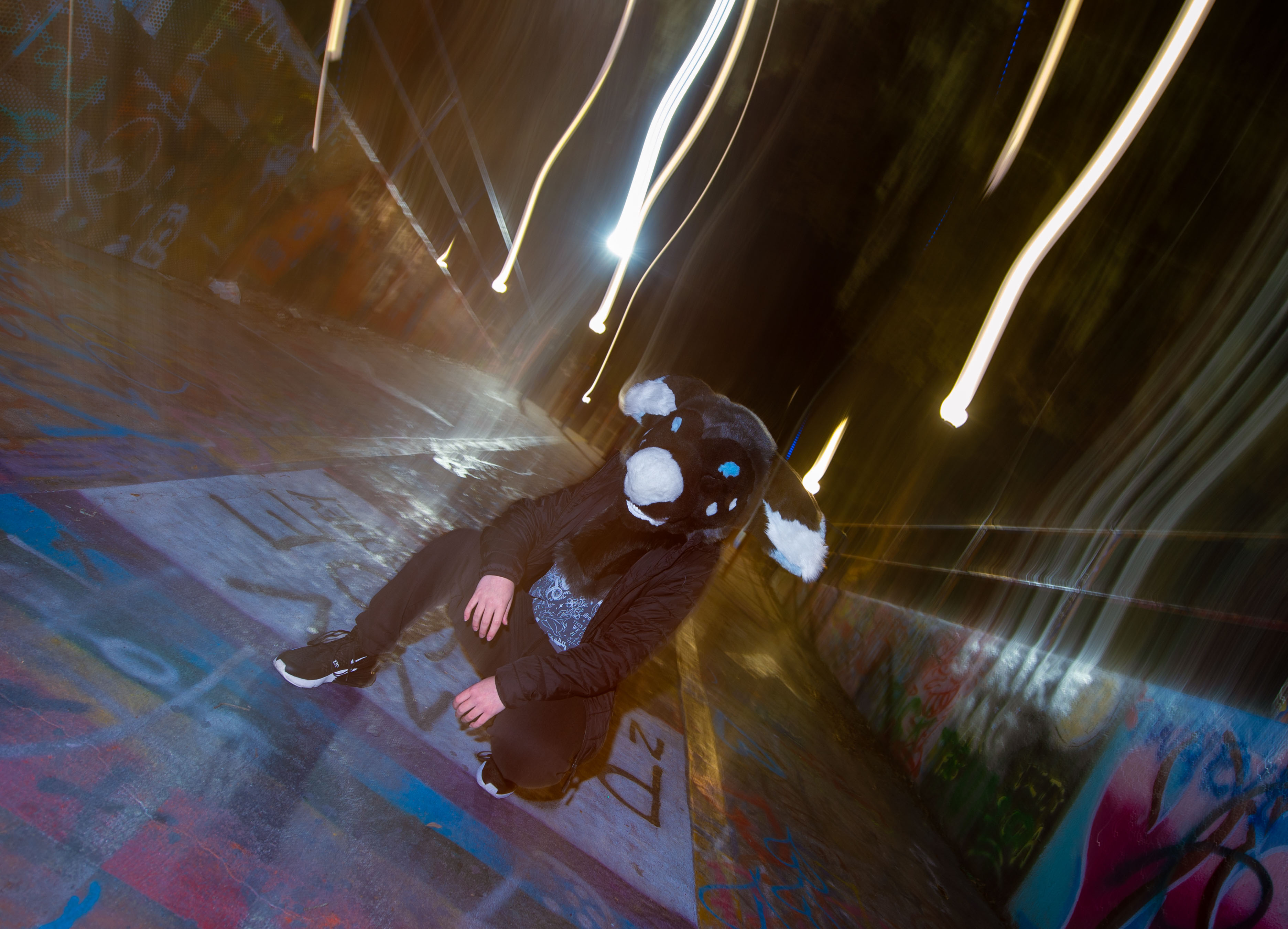 A person in a black, grey, white, and blue fursuit crouching on some heavily-graffitied concrete, with the light painted across the image, as it was taken with a longe exposure