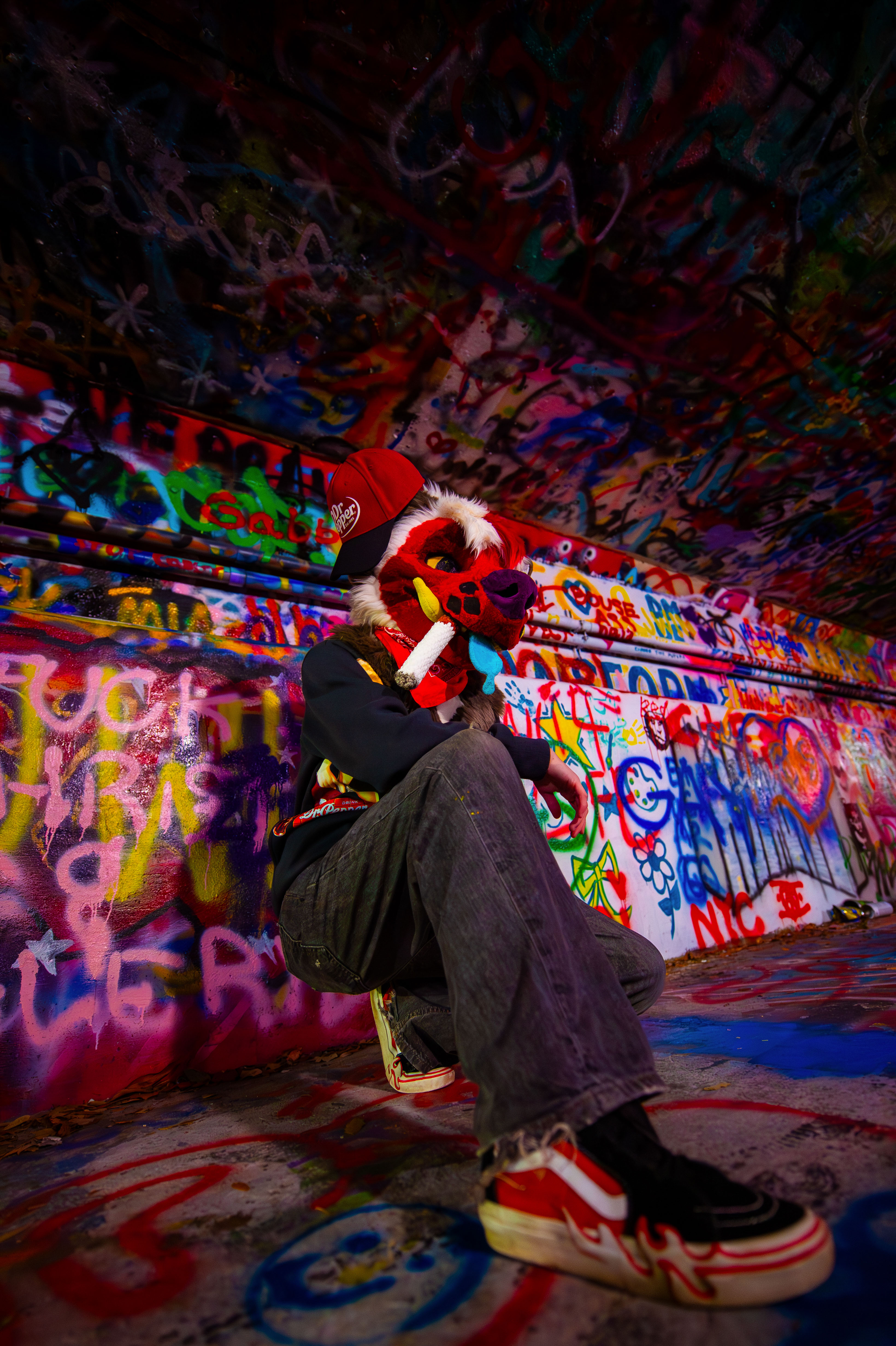 A person in a red fursuit crouching in a heavily graffitied environment/tunnel.
