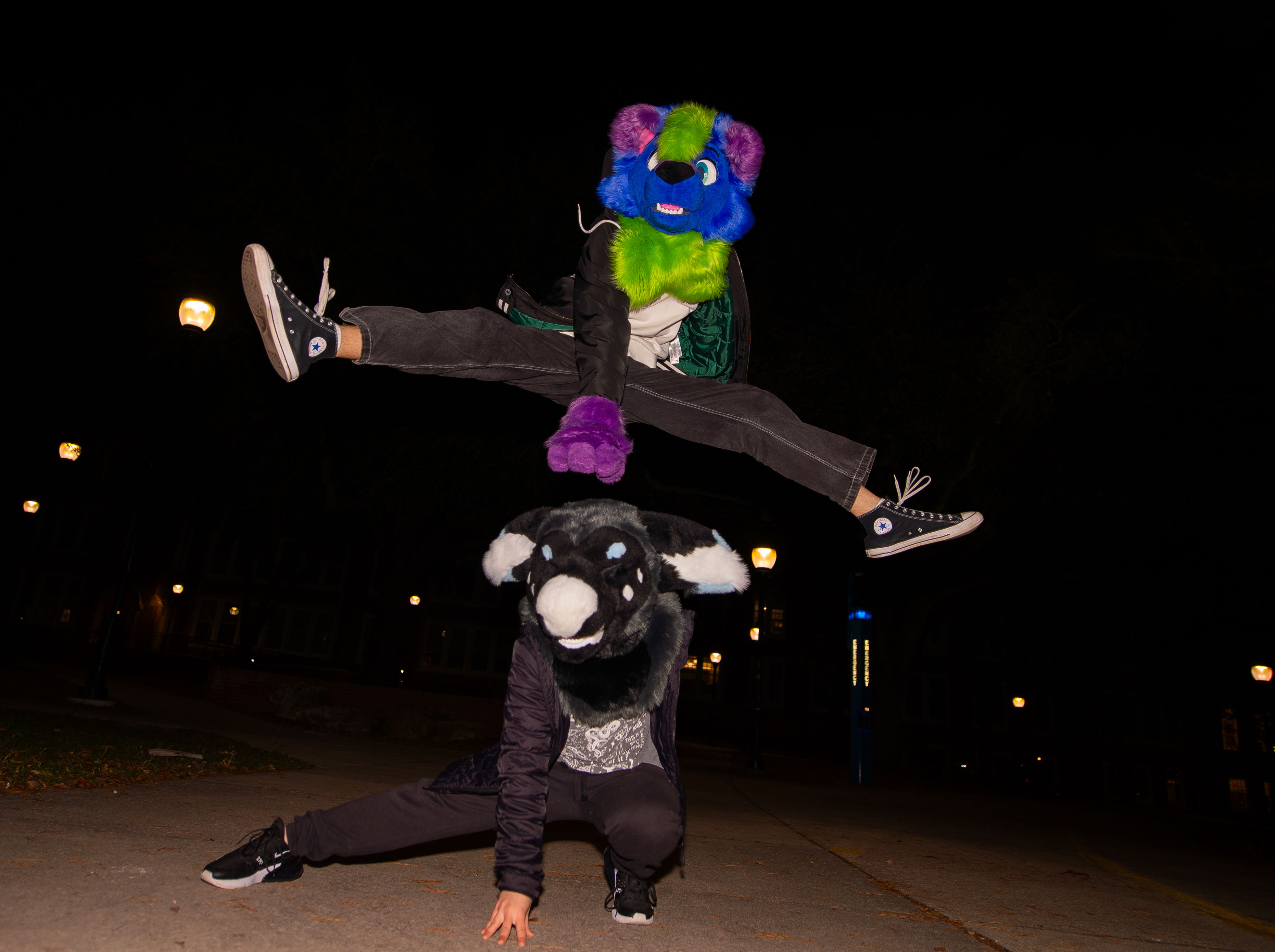 A person in a blue and green fursuit jumping over a person in a black, grey, white, and blue fursuit